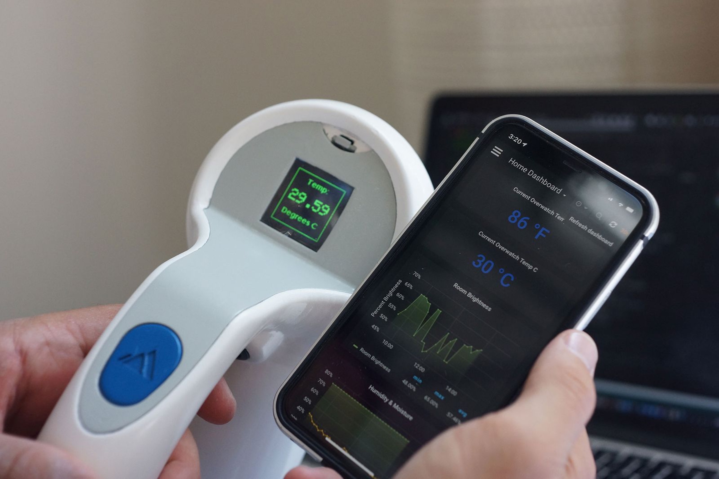 The Theia contactless thermometer next to a phone showing the live temperature dashboard